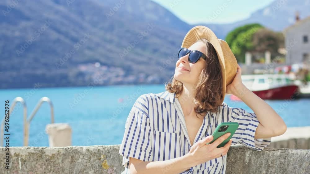 A young Caucasian woman in a hat and sunglasses using her phone while sitting by the sea on a sunny day. A young Caucasian woman in a hat and sunglasses using her phone while sitting by the sea on a sunny day.
