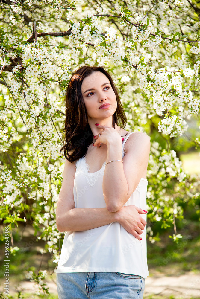 portrait of smiling happy brunette beautiful woman in white T-shirt and pink manicure with white cherry blossoming in the spring sunny day portrait of smiling happy brunette beautiful woman in white T-shirt and pink manicure with white cherry blossoming in the spring sunny day