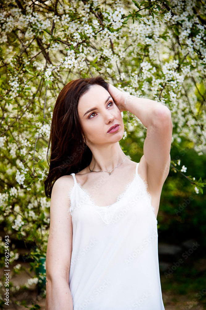 portrait of smiling happy brunette beautiful woman in white T-shirt and pink manicure with white cherry blossoming in the spring sunny day portrait of smiling happy brunette beautiful woman in white T-shirt and pink manicure with white cherry blossoming in the spring sunny day