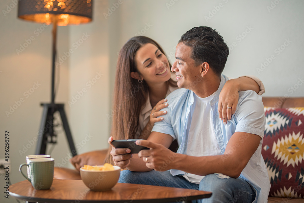 Young couple in real life indoor at home having leisure relax on the sofa and using mobile phone app. Modern young boy and girl living together in apartment. Relationship and dating people lifestyle Young couple in real life indoor at home having leisure relax on the sofa and using mobile phone app. Modern young boy and girl living together in apartment. Relationship and dating people lifestyle
