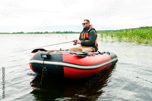 A professional fisherman in a life jacket sits in a boat with a fishing rod. Summer fishing on the pond