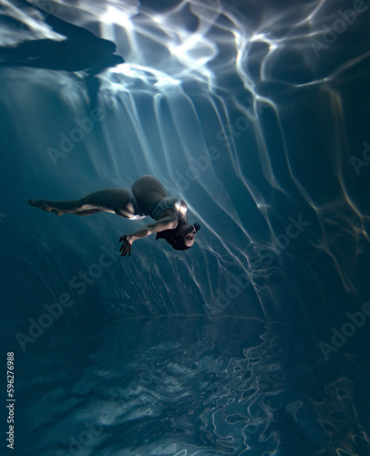 Underwater shoot of beautiful pregnant woman swimming in water through sunbeams.
