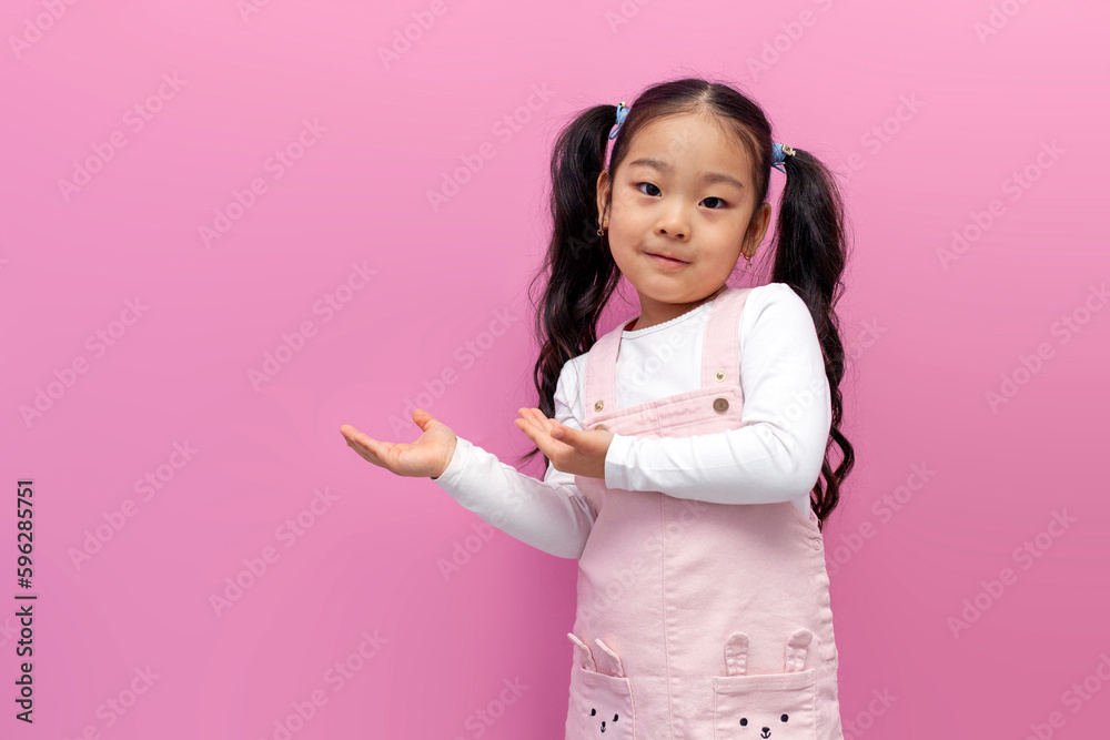 little asian girl in pink sundress with long hair shows her hands to the side on pink isolated background, korean child little asian girl in pink sundress with long hair shows her hands to the side on pink isolated background, korean child
