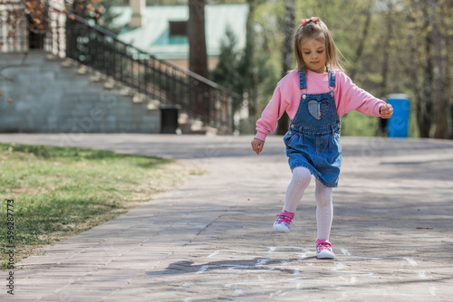 A happy girl in pink clothes draws classics with chalk on the asphalt on the street. Portrait of a little girl playing and jumping hopscotch on a sunny summer day. Creative development of children. © Юлия Клюева