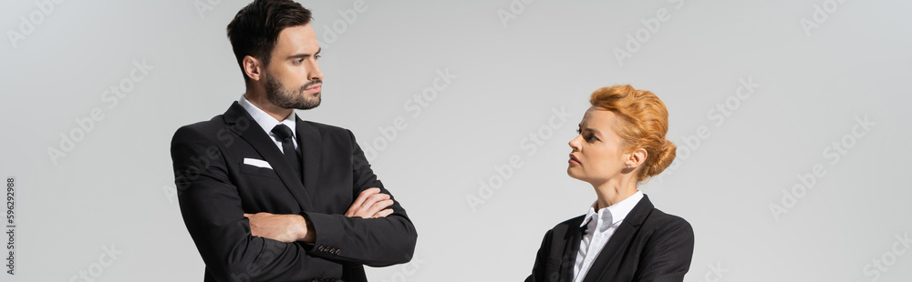 serious and skeptical business people looking at each other while standing with crossed arms isolated on grey, banner.