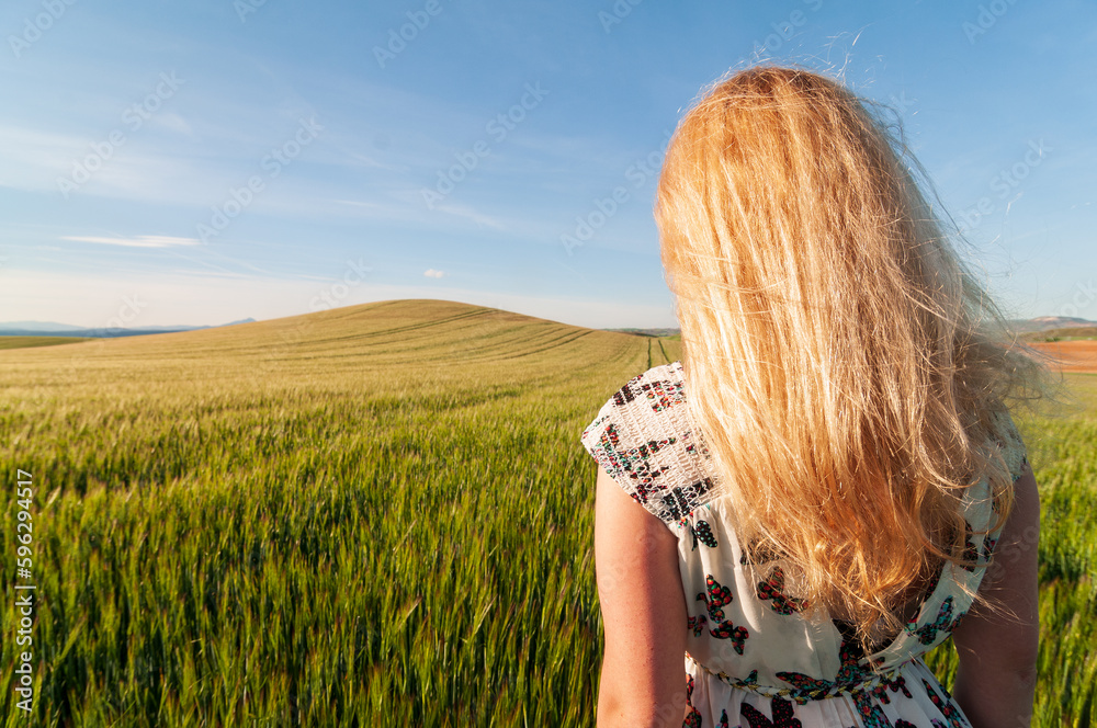 Una mujer rubia mira hacia un campo cultivado de trigo con una suave colina al fondo. Una mujer rubia mira hacia un campo cultivado de trigo con una suave colina al fondo.