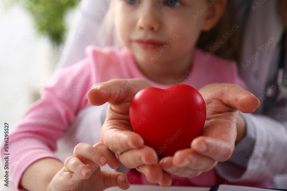 Hands of child and cardiologist doctor with red heart closeup. Treatment of heart disease in children