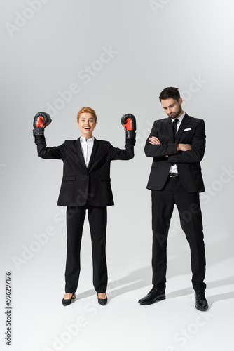 excited redhead businesswoman in boxing gloves showing triumph gesture near sad businessman on grey background.