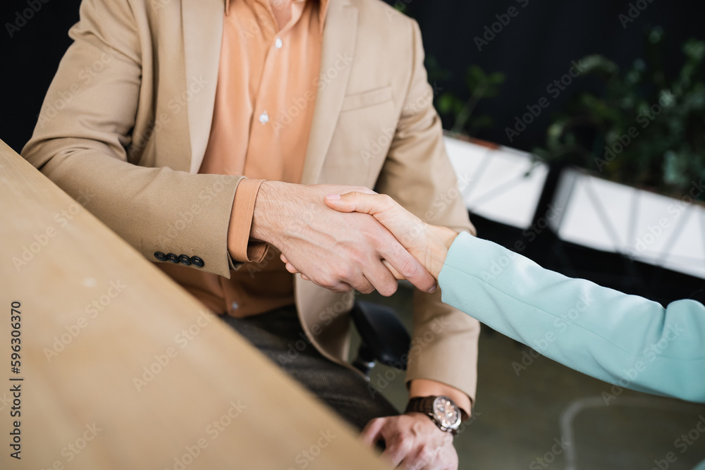 partial view of business partners shaking hands on blurred foreground in office.