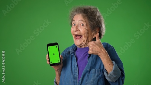 Funny crazy elderly old toothless woman points to phone with green screen tracking points isolated on green screen background with mock up copy space. Hold mobile phone with blank empty screen photo