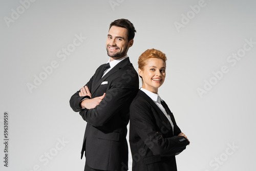 successful corporate managers in black suits standing back to back and smiling at camera isolated on grey.