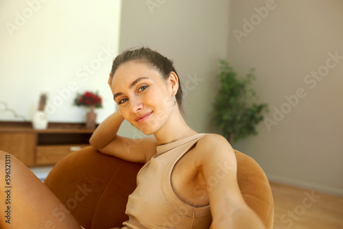 Cute girl taking selfie for dating application profile picture, looking at camera with happy friendly smile, dressed in beige summer tank top, sitting in brown armchair in minimalistic living room