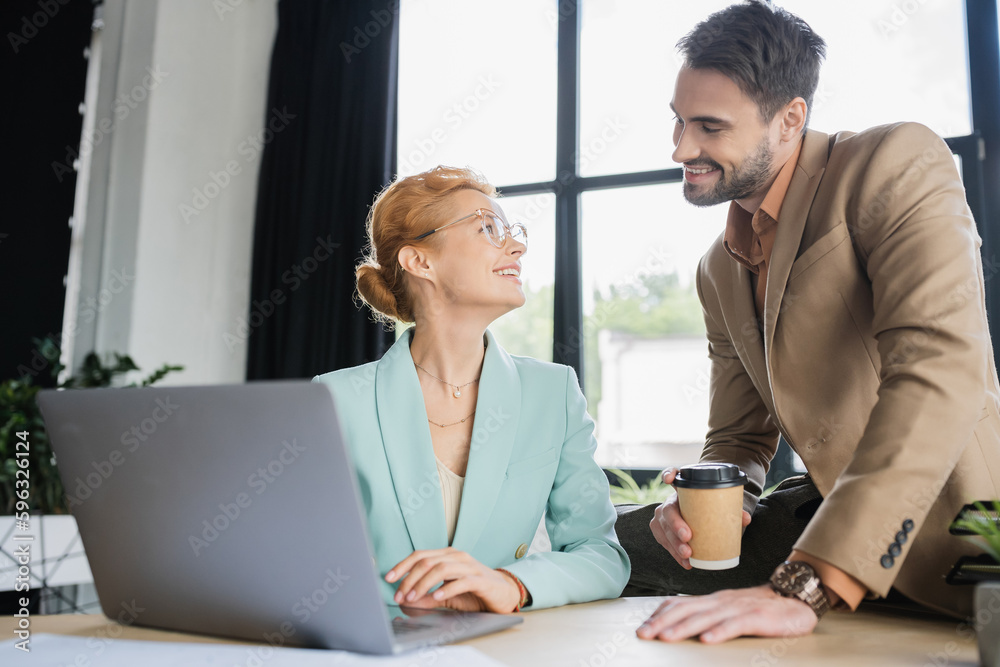 happy businesswoman in eyeglasses working on laptop and looking at bearded colleague with coffee to go in office. happy businesswoman in eyeglasses working on laptop and looking at bearded colleague with coffee to go in office.