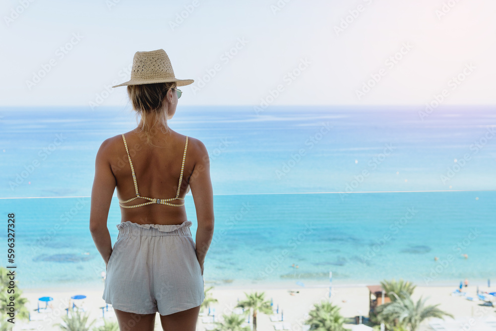 Woman wearing bikini and straw hat enjoy view from beachfront hotel or apartment balcony