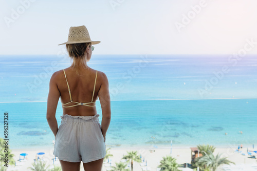 Woman wearing bikini and straw hat enjoy view from beachfront hotel or apartment balcony