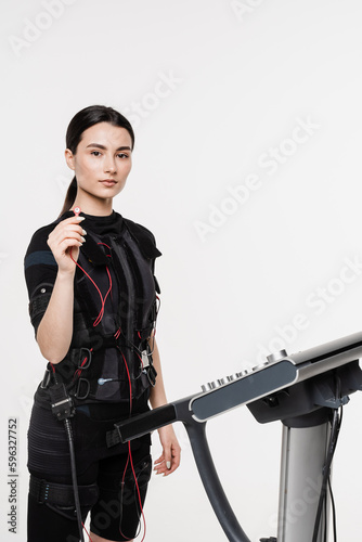 Fit girl in EMS suit with contactor in hand that uses electrical impulses to stimulate muscles on white background. Sport training in electrical muscle stimulation suit.