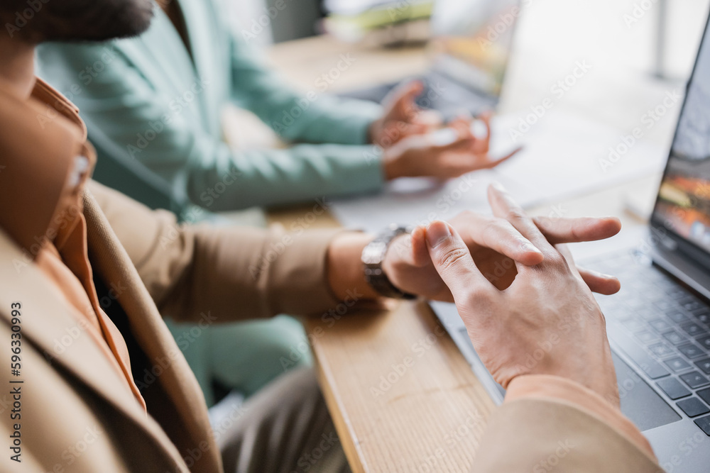 Obraz premium partial view of man sitting with clenched hands near laptop and blurred businesswoman gesturing in office.