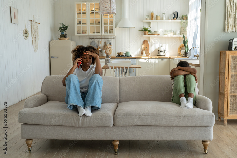 Annoyed angry African American mother sitting with offended upset son on sofa at home, talking on phone, discussing child behavior with spouse. Mom and kid conflict. Parents, children and discipline Annoyed angry African American mother sitting with offended upset son on sofa at home, talking on phone, discussing child behavior with spouse. Mom and kid conflict. Parents, children and discipline