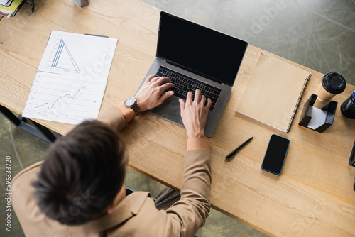 top view of brunette businessman typing on laptop with blank screen near documents and smartphone in office.