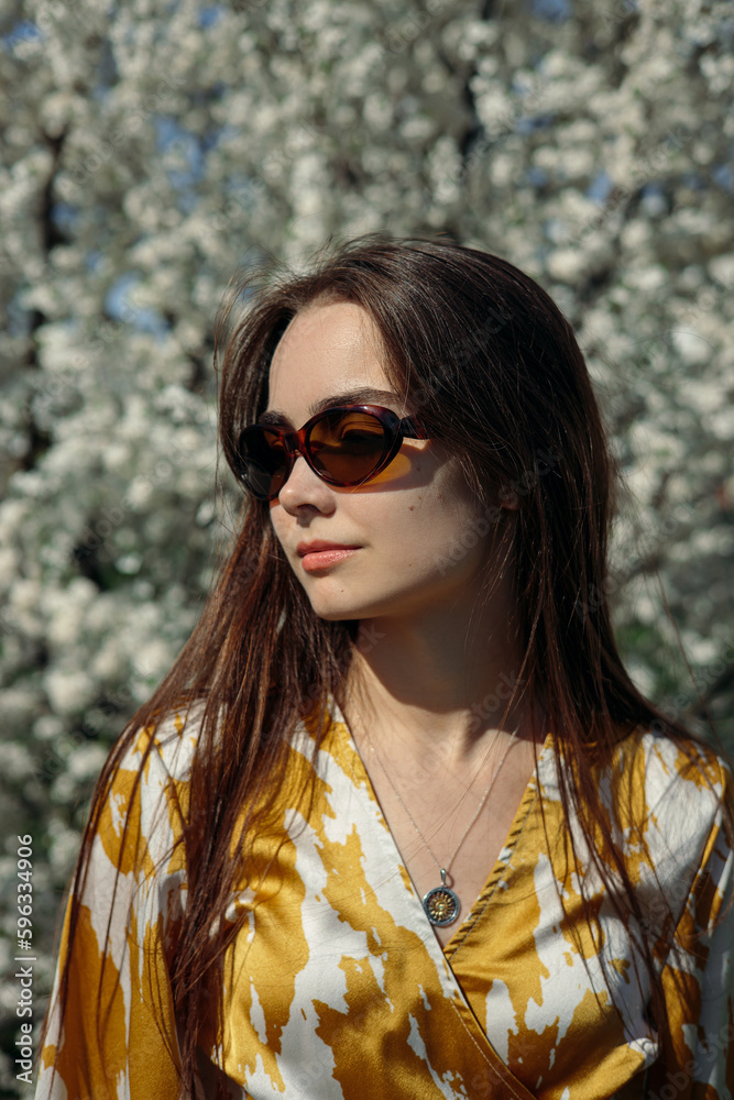 Serene brunette in sunglasses standing in blooming garden with trees in flowers Serene brunette in sunglasses standing in blooming garden with trees in flowers