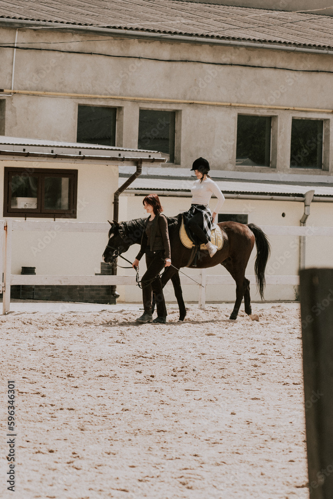 A woman instructor teaching girl how to ride a horse. Female rider practicing on a horseback learning equestrian sport. Active lifestyle and leisure activity concep A woman instructor teaching girl how to ride a horse. Female rider practicing on a horseback learning equestrian sport. Active lifestyle and leisure activity concep