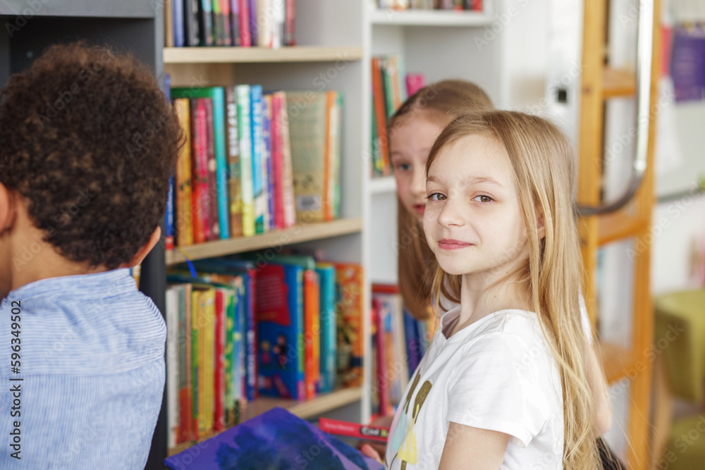 Fototapeta premium Three multiethnic children look for books near bookshelves and read together in school library.