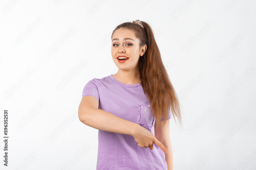 Happy young woman with long hair pointing finger down and smiling, showing advertisement, product below, promo offer, standing in casual t shirt over white background Happy young woman with long hair pointing finger down and smiling, showing advertisement, product below, promo offer, standing in casual t shirt over white background