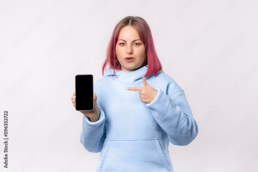 Surprised lovely woman with pink hair in a blue hoodie shows a mobile phone with a blank screen. Young female looking amazed by promotion offer, stands over white background Surprised lovely woman with pink hair in a blue hoodie shows a mobile phone with a blank screen. Young female looking amazed by promotion offer, stands over white background