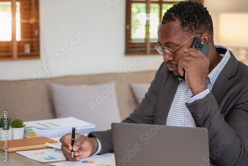 An African American businessman in a black suit uses a mobile phone to communicate with clients and a laptop to view company profits. to build confidence for customers to come and invest in the office