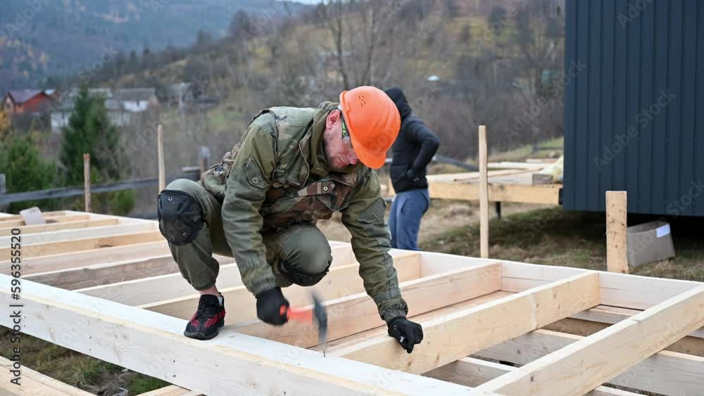 Man worker building wooden frame house on pile foundation. Carpenter hammering nail into wooden joist, using hammer. Carpentry concept. Man worker building wooden frame house on pile foundation. Carpenter hammering nail into wooden joist, using hammer. Carpentry concept.