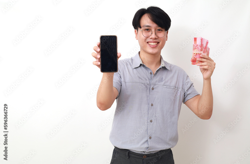 Happy asian man holding Indonesian banknotes while showing blank cell phone display. Isolated Happy asian man holding Indonesian banknotes while showing blank cell phone display. Isolated