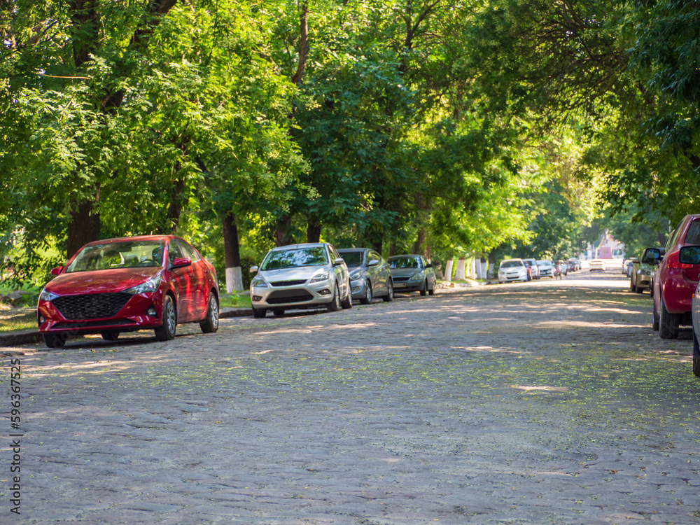 Parked cars in the shade of trees on a cobblestone street. City Alley