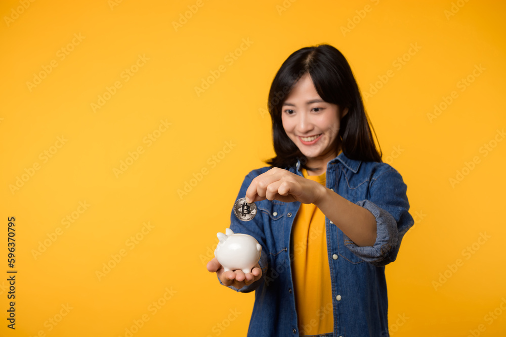 Happy young asian woman wearing yellow t-shirt denim shirt saving digital crypto currency to piggy bank isolated on yellow background. Digital money saving investment concept. Happy young asian woman wearing yellow t-shirt denim shirt saving digital crypto currency to piggy bank isolated on yellow background. Digital money saving investment concept.