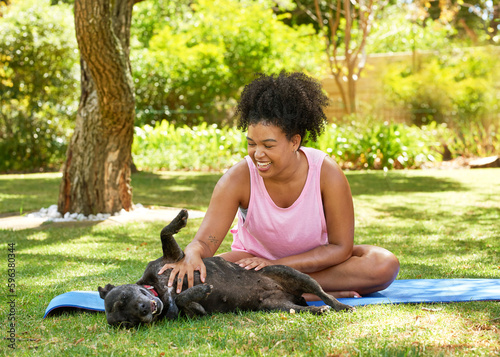 Young multi-ethnic woman gives her dog a tummy tickle on yoga mat in park photo