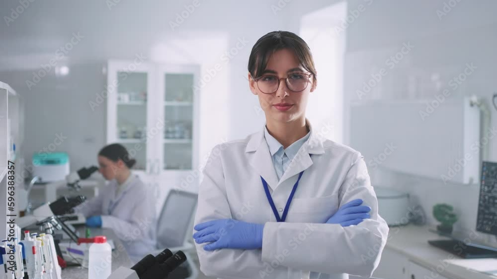 Portrait of female scientist with arms crossed looking at camera. Young smiling professional in white lab coat on blurred background of laboratory room. Daytime. Lifestyle. Indoors Portrait of female scientist with arms crossed looking at camera. Young smiling professional in white lab coat on blurred background of laboratory room. Daytime. Lifestyle. Indoors