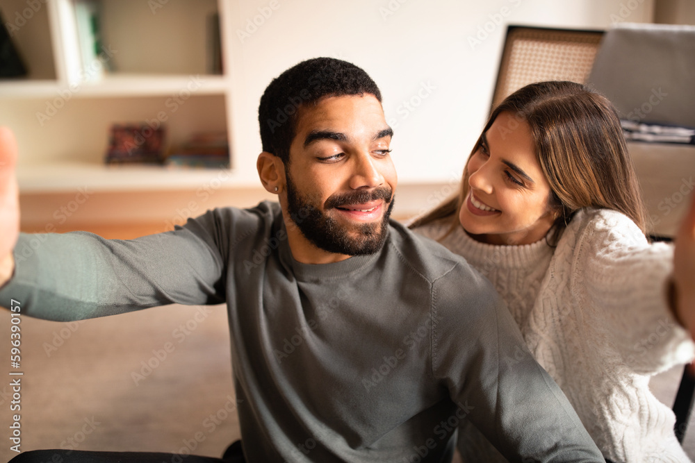 Glad millennial international husband and wife have fun, take selfie on phone together in living room interior Glad millennial international husband and wife have fun, take selfie on phone together in living room interior