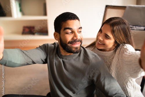 Glad millennial international husband and wife have fun, take selfie on phone together in living room interior