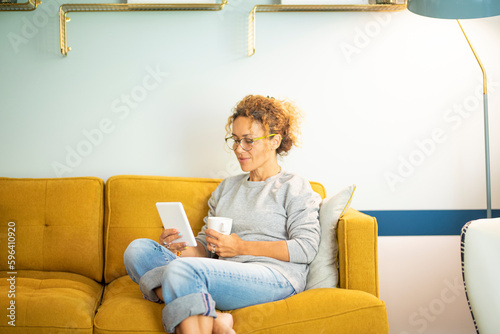 Smart casual woman middle age relaxing at home sitting on yellow sofa and using ereader to read an ebook enjoying indoor leisure activity alone. Sunday time lifestyle. People and technology online photo