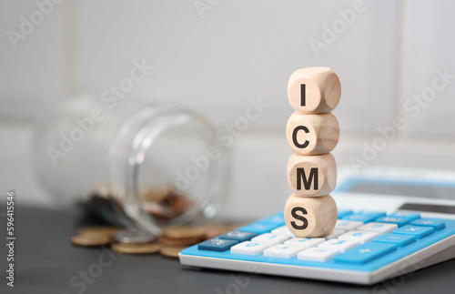 A sigla ICMS escrita em dados de madeira que estão sobre uma calculadora. Um pote de vidro com moedas do Brasil na composição. photo