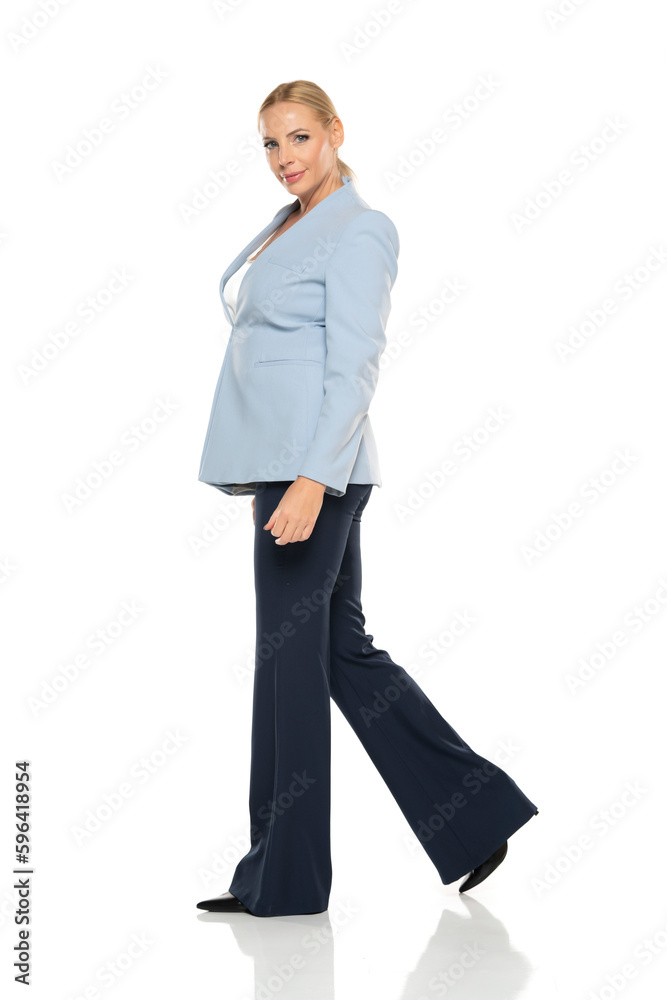Middle aged senior smiling woman wearing blue jacket and black trousers walking on a white background. Profile view Middle aged senior smiling woman wearing blue jacket and black trousers walking on a white background. Profile view