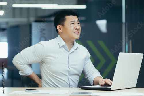 Tired young asian man working overtime in the office on a project, feeling severe back pain from sitting, holding and massaging his back with his hand.