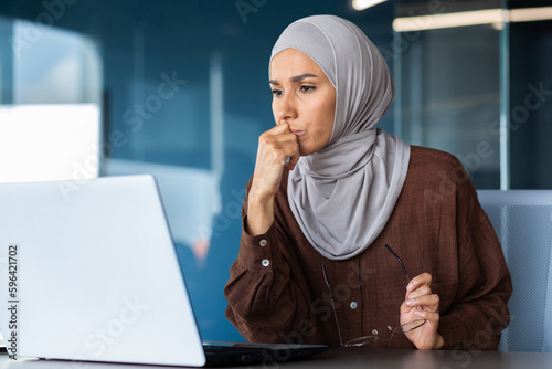 Fotografía Portrait of a serious young woman in a hijab sitting in front of a laptop, holding glasses, thoughtfully and intently looking at the monitor