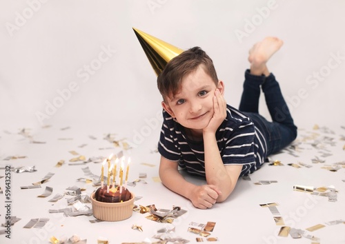 A boy of 7 years old in a striped T-shirt and jeans in a gold cap celebrates his birthday. He holds a donut with lit candles in his hands on a white background. The concept of children's birthday, chi