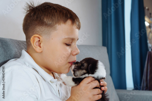 Teenage boy is sitting on couch holding Welsh corgi puppy in his hands and gently kissing him on nose. Caring for and caring for pets. Love and friendship. Positive. Happy childhood.