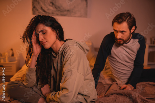 Upset woman sitting near blurred boyfriend talking on bed in evening.