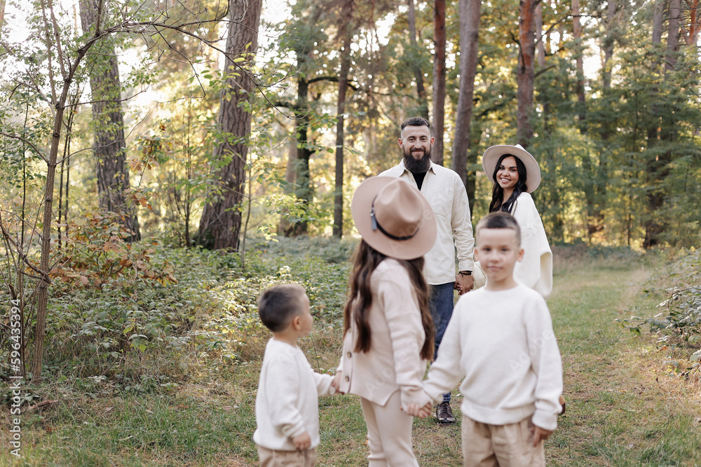 Young mother, father with daughter and sons are walking, having fun in autumn forest. Family holding hands enjoying time together at background of trees. Happy parenthood and childhood concept.