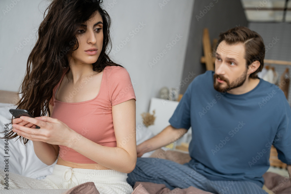 Brunette woman hiding smartphone near blurred angry boyfriend on bed. Brunette woman hiding smartphone near blurred angry boyfriend on bed.