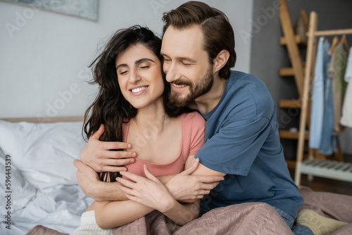 Overjoyed man hugging brunette girlfriend in pajama on bed at home.