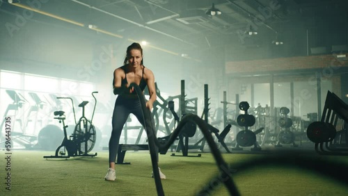 Well-built cute woman exercises on crossfit battle rope in gym. Spacious room, power equipment, cardio machines, large mirror in the background. High quality 4k footage