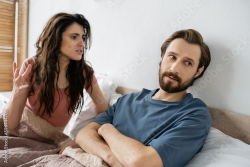 Angry woman talking to bearded boyfriend crossing arms on bed at home.